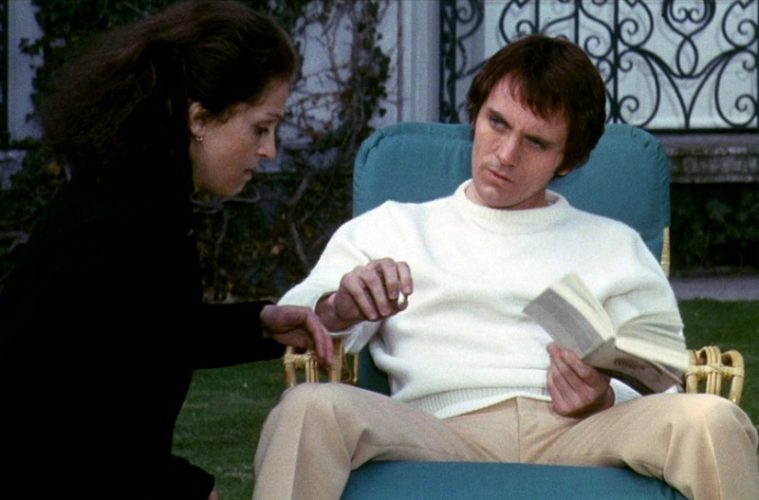 Lucia talking to The Visitor while he sits in a chair smoking a cigarette. Lucia has a concerned look on her face.