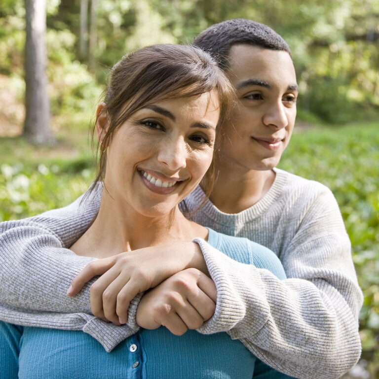 A latin mom smiles as her son has his arms around her and smiles while they are outdoors.
