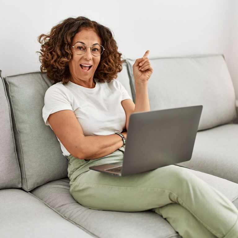 A middle aged latin woman smiles while working on her laptop on her couch with a finger pointing outwards.