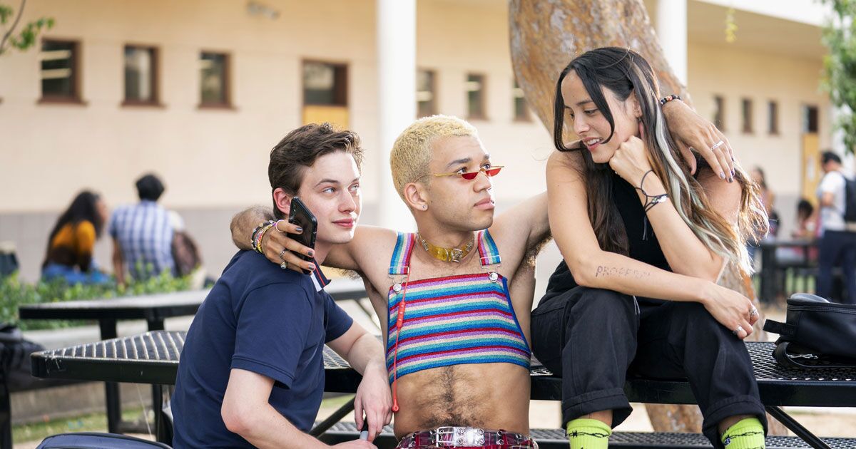 Chester con sus brazos alrededor de Nathan y Riley, quienes est&aacute;n sentados a su lado en una mesa para almorzar al aire libre y le sonr&iacute;en.