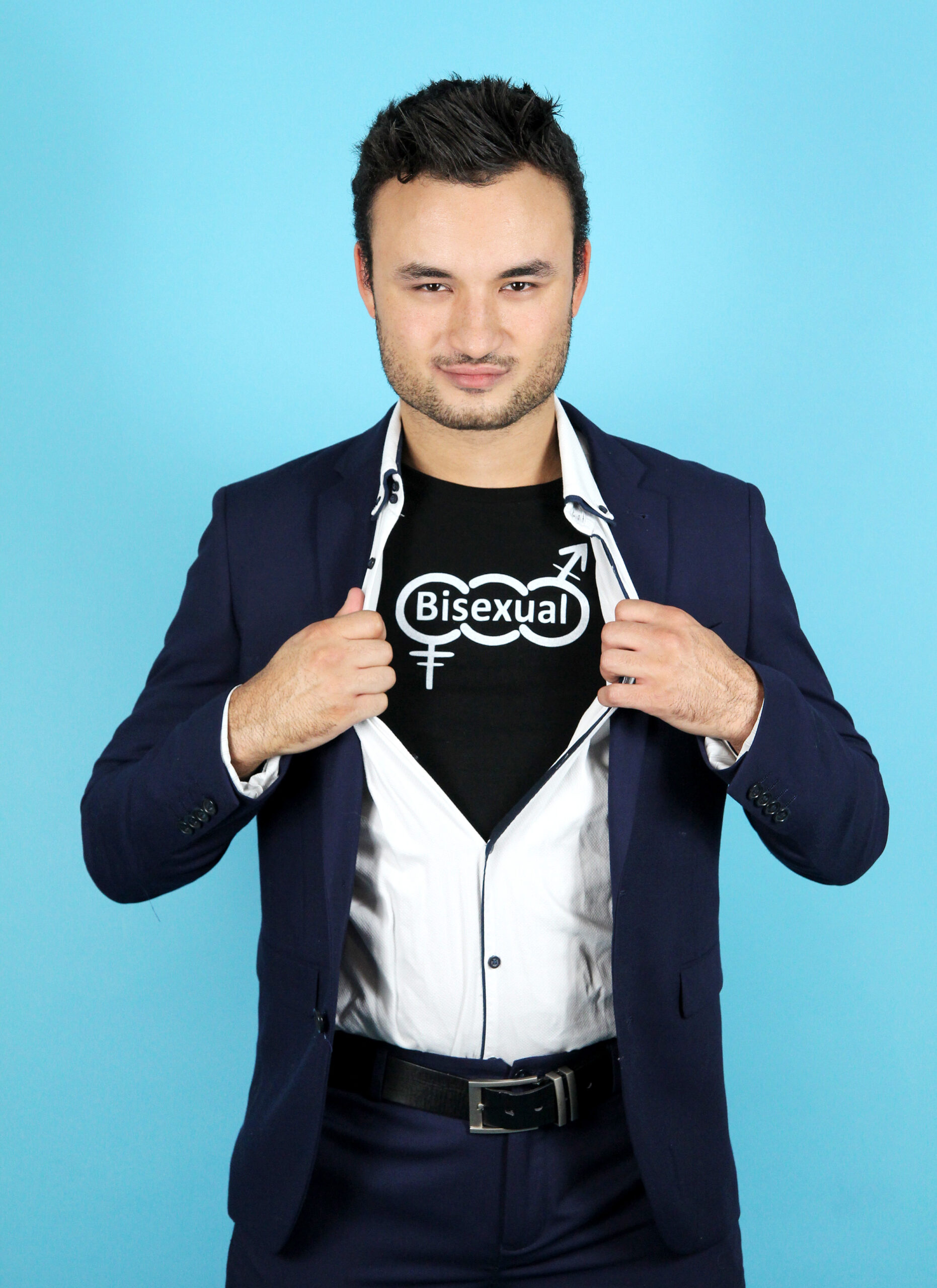 Lewis Oakley vistiendo un blazer y abriendo la parte superior de su camisa para exponer una camisa con las palabras bisexual en ella.