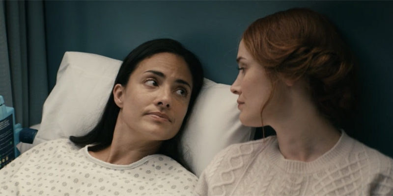 Queen Maeve looking at Elena who is laying on a hospital bed with a serious look on her face.