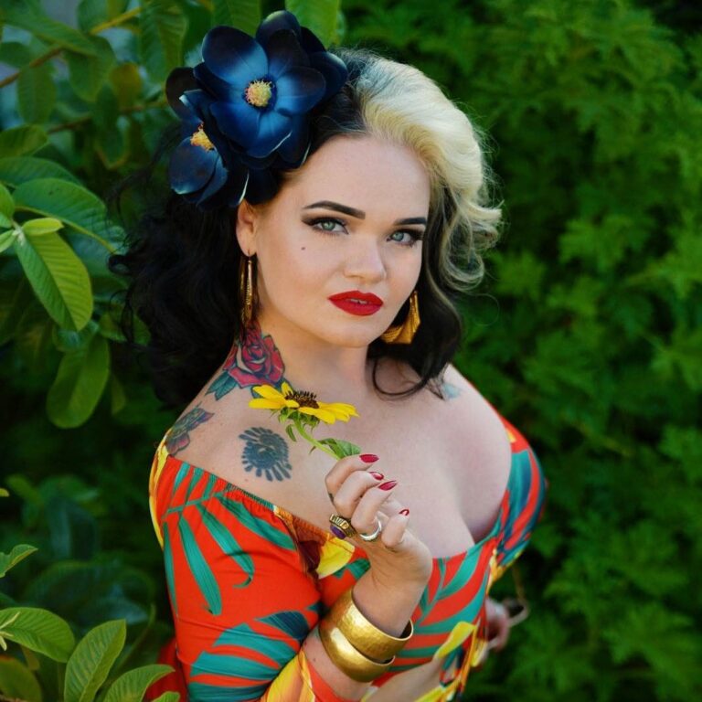 Molly Moonstone in front of a garden wearing a colorful dress and posing with a flower.