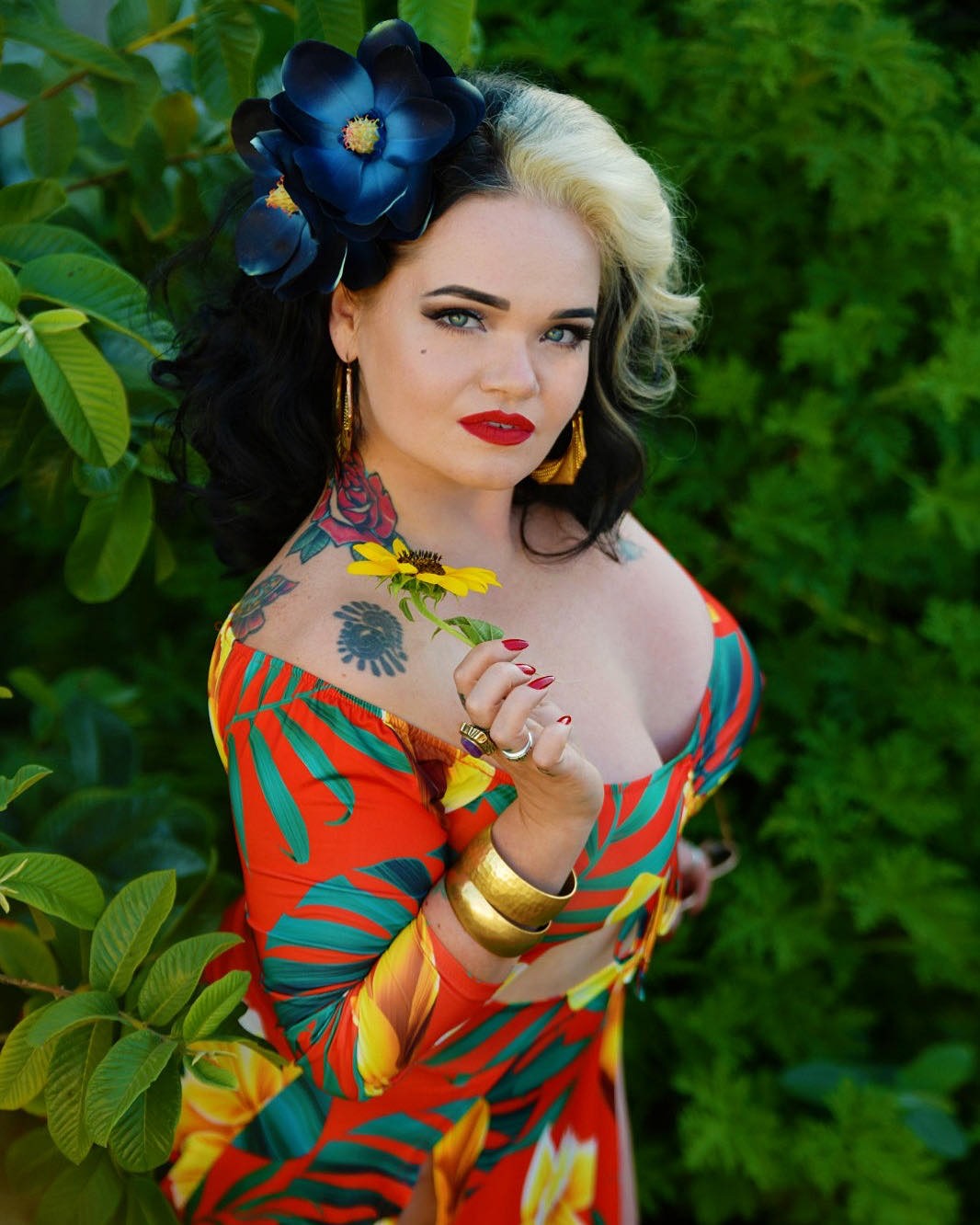 Molly Moonstone in front of a garden wearing a colorful dress and posing with a flower.