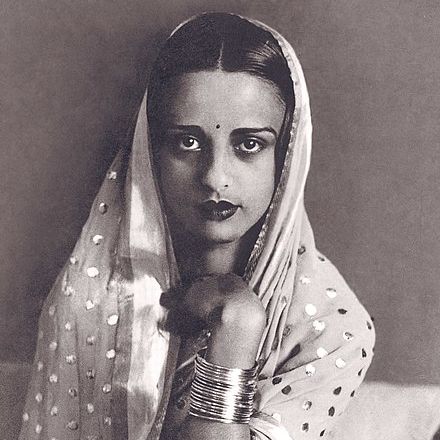 Black and white portrait of Amrita. She has on a silk head covering with many bracelets. She smiles calmly in front of her a hand on her neck.