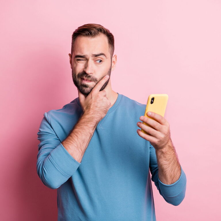 A man looks at his phone with one eye while contemplating, holding a hand to his chin against a pink background.