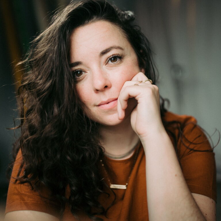 Headshot of Becca with her hair to one side and a hand on her chin smiling.