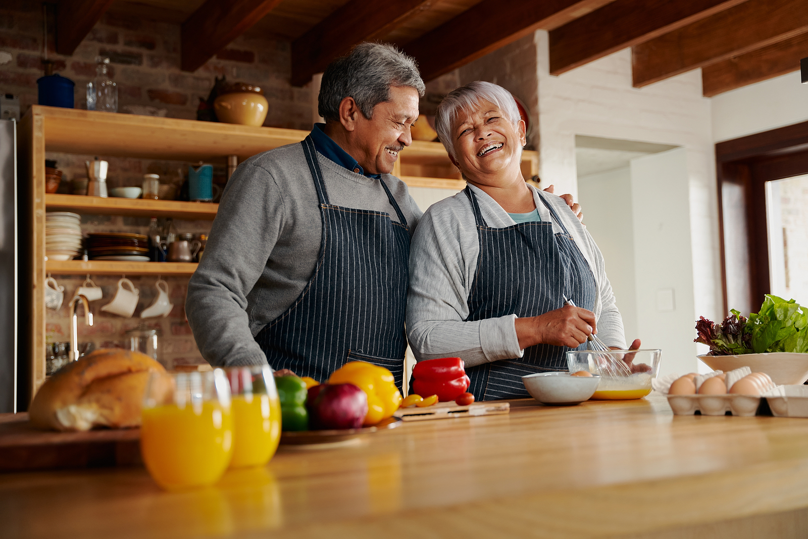 Una pareja casada birracial se r&iacute;e mientras prepara el desayuno en su cocina. El marido tiene su mano en el hombro de su esposa.