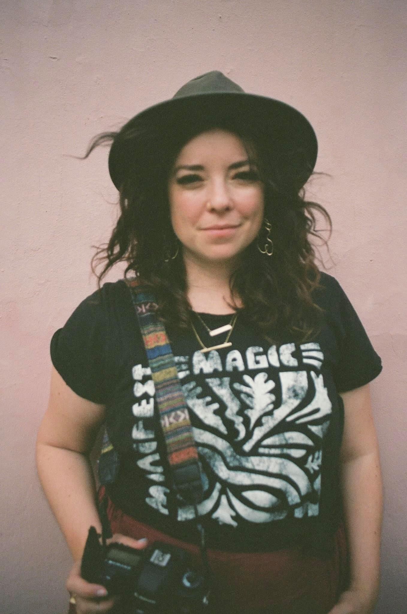 Rebeca wearing a stylish outfit with her camera on her side smirking. The photograph has a grainy aesthetic.