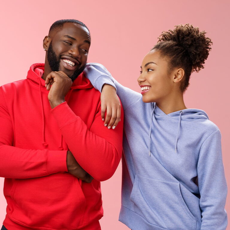 An attractive man and woman smile to eachother. The woman has her arm over his shoulder while his arms are crossed.
