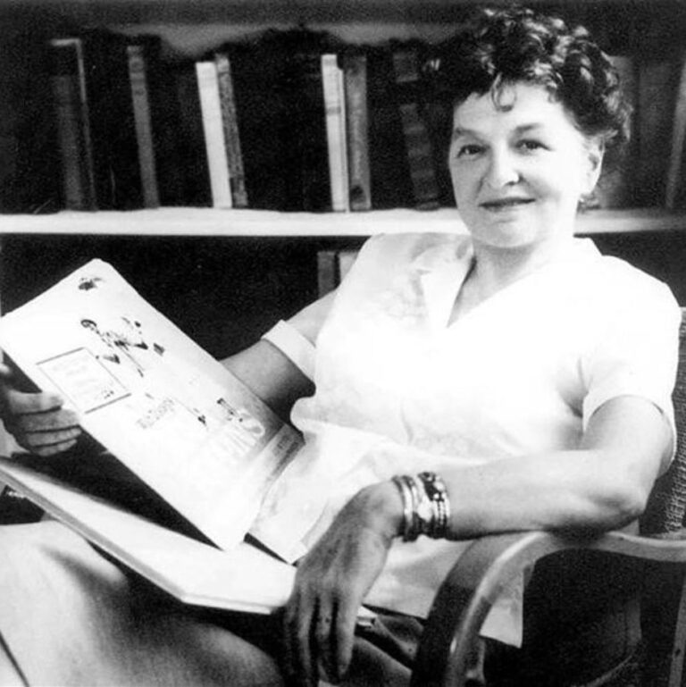 Black and white image of Travers sitting on a wodden chair in front of a bookshelf, smiling calmly with a book on her lap.