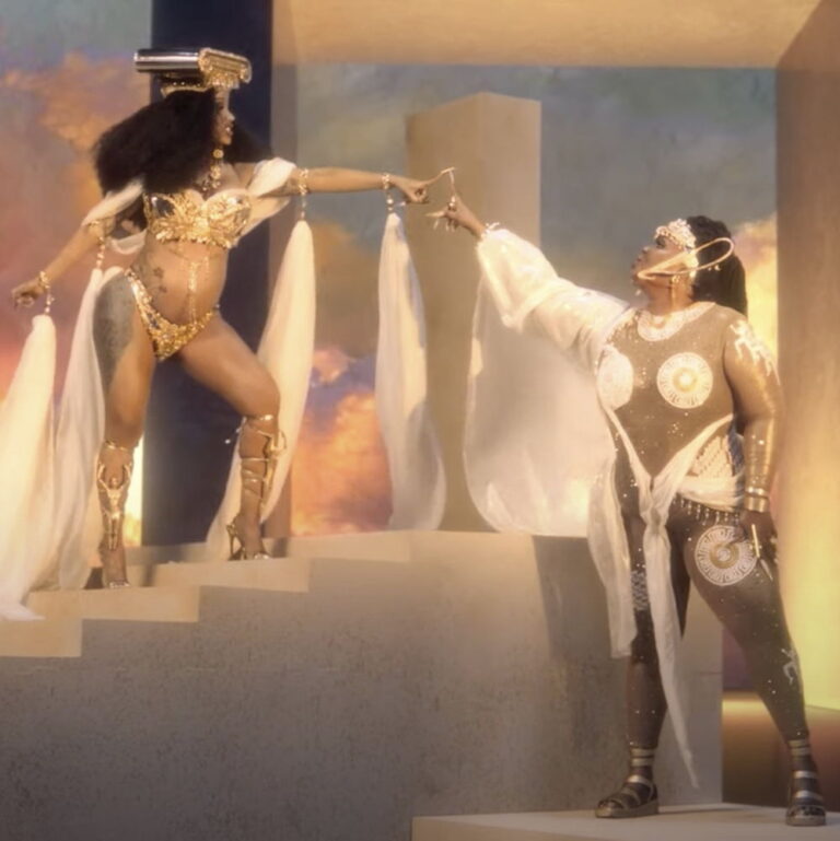 Lizzo and Cardi B from Rumors. Both are waring gowns and gold, touching finger tips.