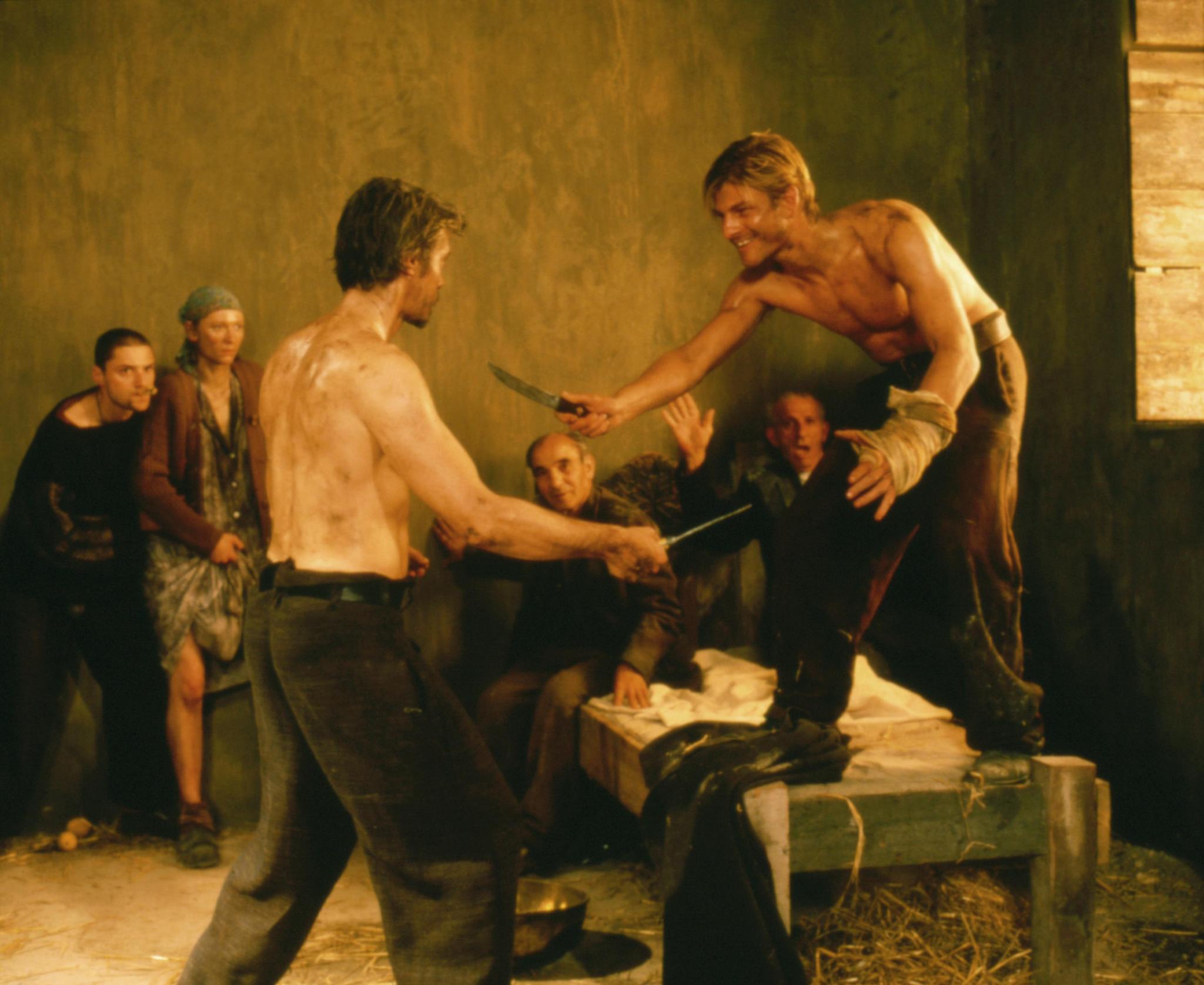 Ranuccio and Caravaggio in the middle of play fighting with knives while others watch. They are shirtless and standing on a frame.