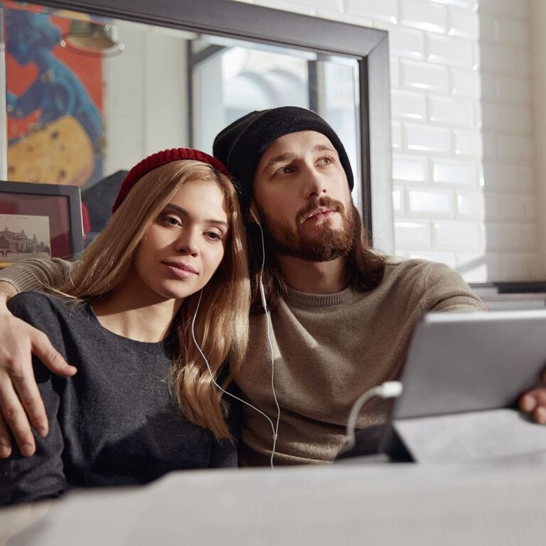 Young thoughtful european couple in headphones watching something on digital tablet in modern cafe.