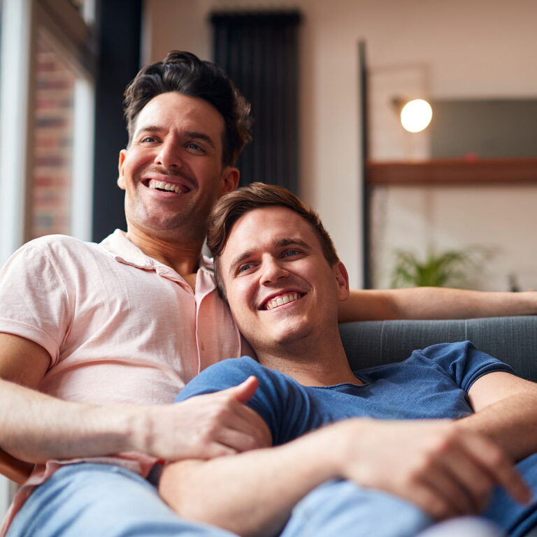 An attractive gay couple is cuddling on the couch together smiling watching tv.
