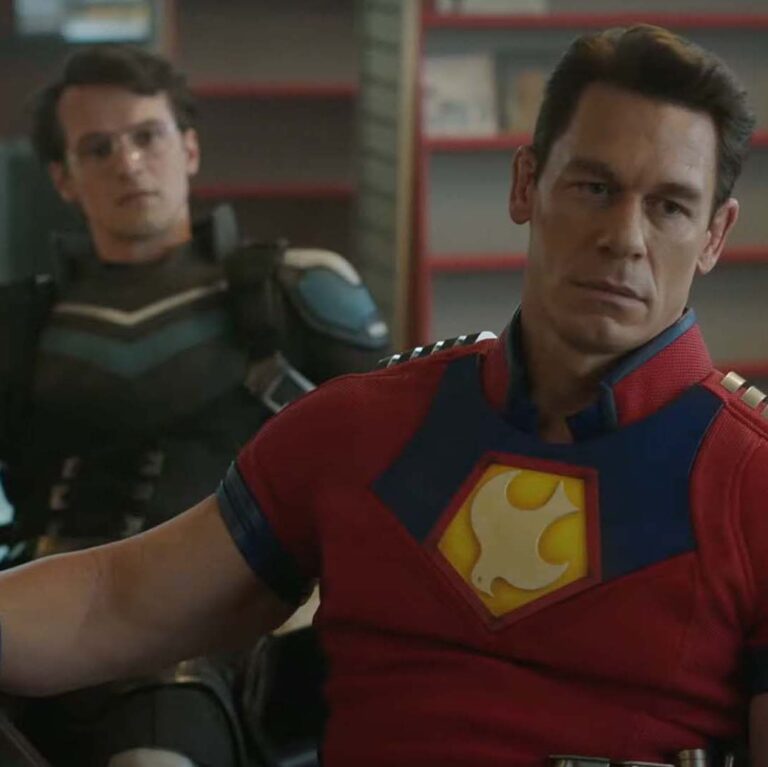 Peacemaker and Vigilante sitting at a table together looking at someone out of frame with serious expresions.