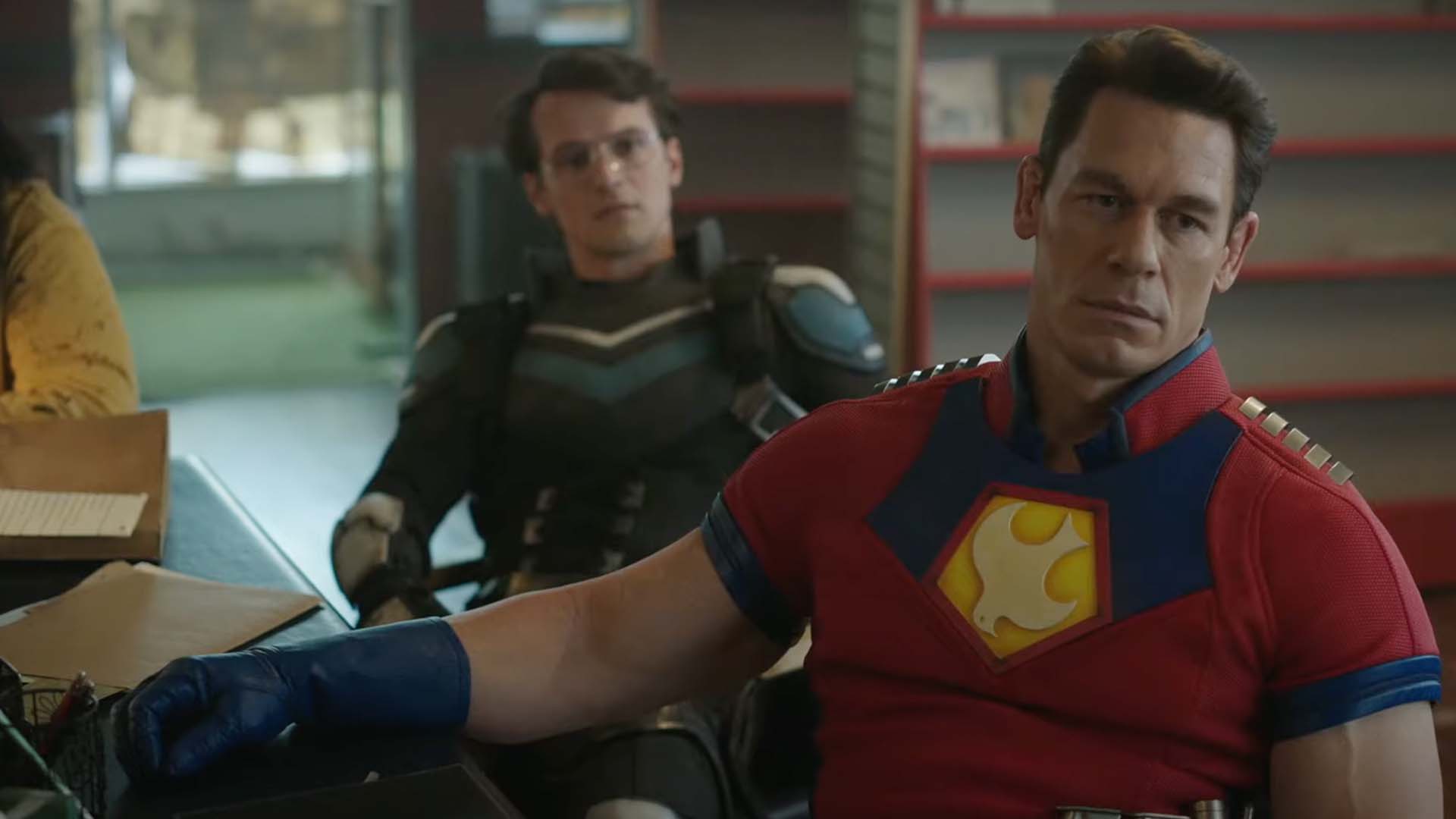 Peacemaker and Vigilante sitting at a table together looking at someone out of frame with serious expresions.