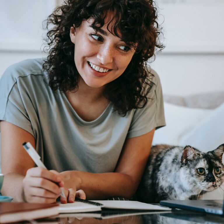 A woman smiles while writing next to her computer. There is a small cat next to her watching her work.