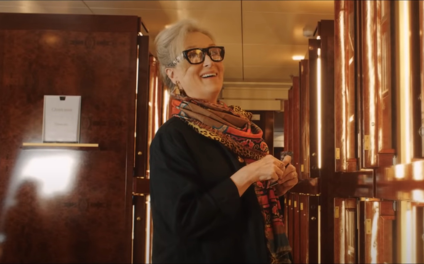 Alice standing in the hallway with her hands held together smiling and talking to someone out of frame.