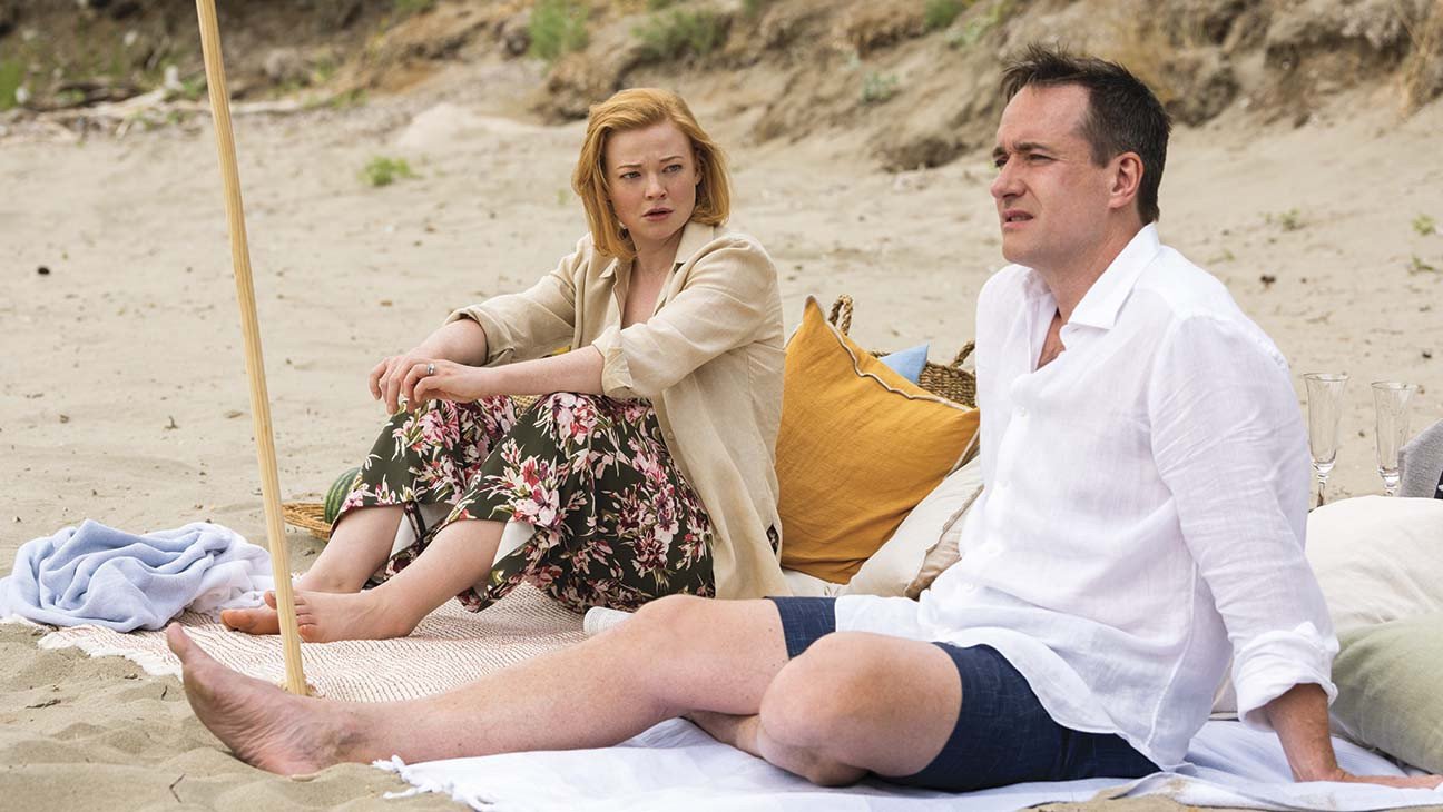 Tom and Shiv sitting together at the beach having a conversation with serious expressions.