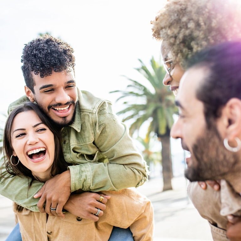 Two pairs of attractive couples laugh together while they carry their partners on their backs outdoors.