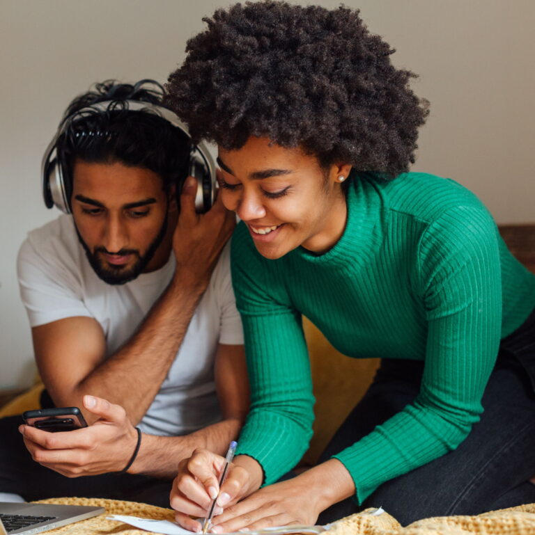 An attractive man and woman sit closet together on a bed while listening to music and writing things down.
