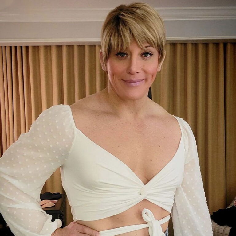 Janae smiling and posing with an arm on her waist wearing a white low cut dress.