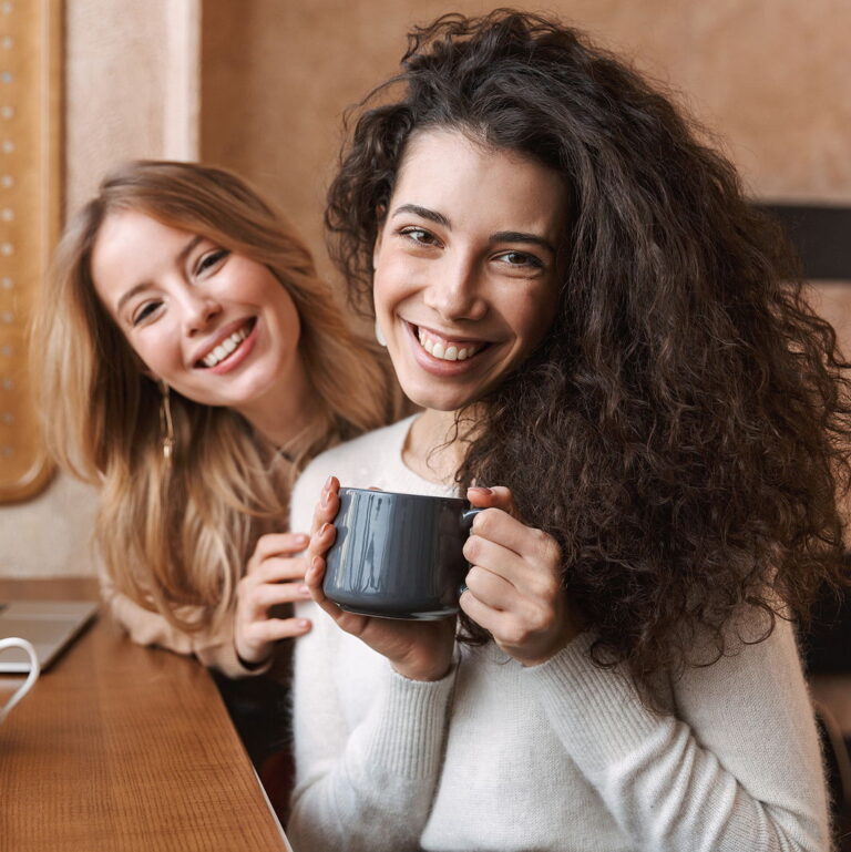 Two cheerful young girlfriends sitting at the cafe indoors, having cup of coffee