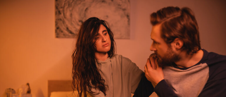 Dissatisfied woman looking at blurred boyfriend sitting on bed at night.